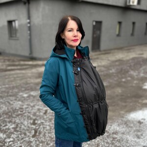 May include: Teal blue hooded jacket with a black baby carrier attachment. The jacket has a drawstring waist, front pockets, and a black zipper. The baby carrier attachment is black and has a drawstring closure.