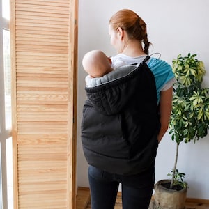 May include: A black quilted baby carrier cover featuring a hood and a soft grey interior lining. The cover is designed to be worn over a baby carrier, providing a layer of warmth with its padded construction and secure fit around the infant.