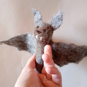 Può includere: Un pipistrello in feltro fatto a mano con le ali spiegate. Il pipistrello presenta un corpo marrone, ali grigie e grandi orecchie grigie. Ha piccole zanne bianche e macchie scure sul viso. Un oggetto artigianale stravagante.
