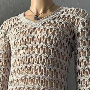May include: Two open-weave crochet tops on mannequins. One is a sleeveless tank in shades of orange and yellow. The other is a long-sleeved, V-neck sweater in a light beige color. Both garments have a distinctive, airy design.