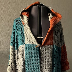 May include: A hooded crocheted cardigan with a patchwork design in shades of gray, teal, and orange. The cardigan has a button closure and is made with a chunky yarn.