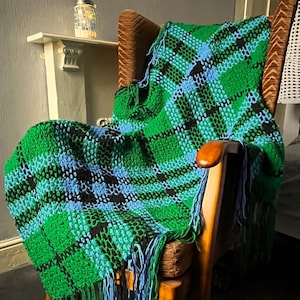 May include: A green, blue, and black plaid crocheted throw blanket with fringe on a wooden armchair.