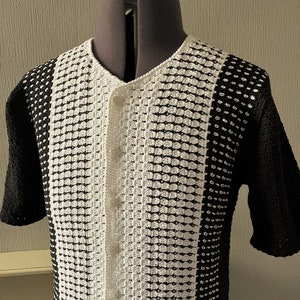 Puede incluir: Un cárdigan de crochet blanco y negro con cierre de botones. El cárdigan tiene mangas cortas y un patrón texturizado.