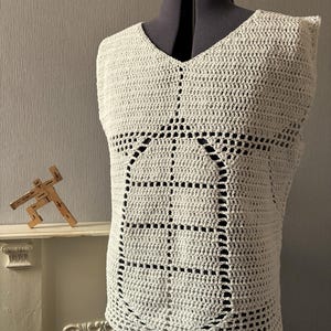 May include: A sleeveless, off-white crochet top featuring a central abdominal muscle pattern created with open-work stitches. The garment has a V-neckline and a structured, boxy fit, displayed on a dark mannequin torso to highlight the texture.