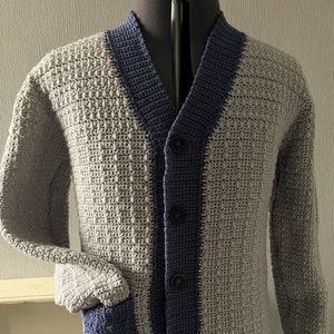 May include: A grey crocheted cardigan with a blue trim and buttons. The cardigan has a pocket on the left side.