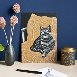 May include: A black and white cat illustration on a card, seated on a gold-colored chair. The card is propped up on a desk with a black vase holding dried flowers, a small tin, and a pen. The background is a dark blue wall.