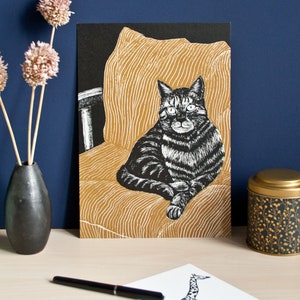 May include: A black and white print of a tabby cat sitting on a gold-colored, striped cushion. The print is displayed on a black background. A small drawing of a giraffe, a black pen, a vase with dried flowers, and a decorative tin are also visible.