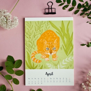May include: A rectangular wall calendar page for the month of April featuring a central illustration of an orange tabby cat crouching in green grass and foliage. The calendar grid is printed in black text below the cat illustration on a white background.