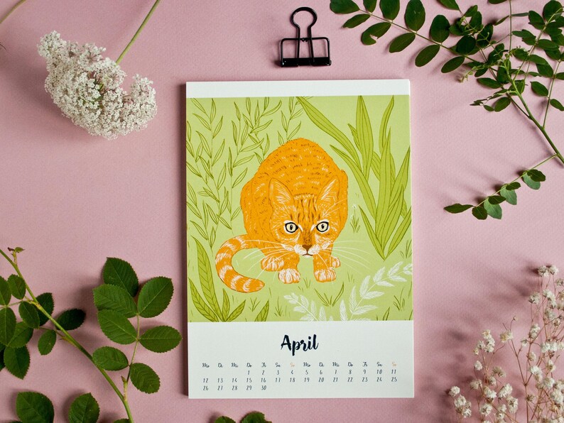 May include: A rectangular wall calendar page for the month of April featuring a central illustration of an orange tabby cat crouching in green grass and foliage. The calendar grid is printed in black text below the cat illustration on a white background.