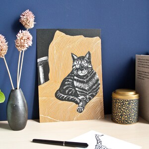 May include: A black and white cat art print. The cat is depicted sitting on a yellow chair. The print is displayed on a desk with a black vase holding dried flowers, a decorative tin, and books. A pen and a small giraffe print are also on the desk.