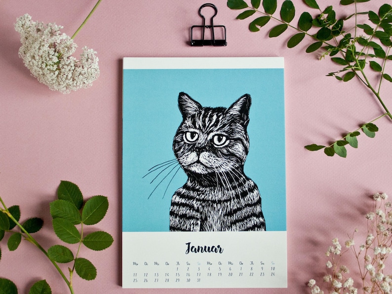 May include: A wall calendar page for the month of January featuring a black and white ink-style illustration of a tabby cat against a solid light blue background. The calendar grid is printed in black text on a white base at the bottom of the page.