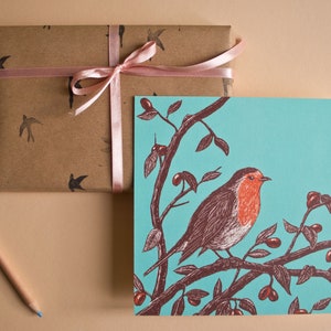May include: A teal blue card with a robin perched on a branch with red berries. The robin is brown and red with a white breast. The card is wrapped in brown paper with a pink ribbon.