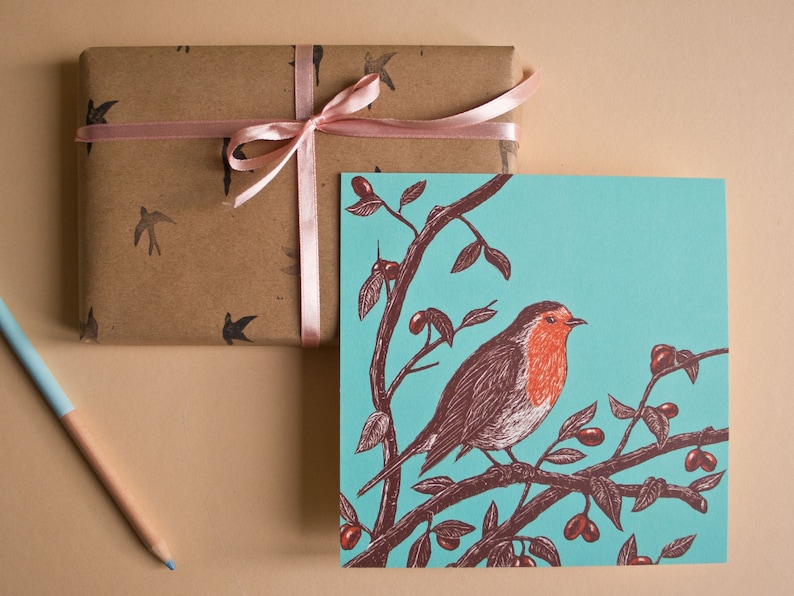 May include: A teal blue card with a robin perched on a branch with red berries. The robin is brown and red with a white breast. The card is wrapped in brown paper with a pink ribbon.