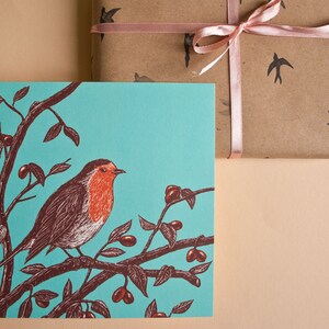 May include: A turquoise blue greeting card with a robin perched on a branch with red berries. The robin is brown and white with a red breast.