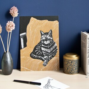 May include: A black and white cat art print on a black background, set against a gold-colored chair illustration. The print is displayed on a light-colored surface, with a black pen and a giraffe sketch. A black vase with dried flowers and a decorative tin are also present.