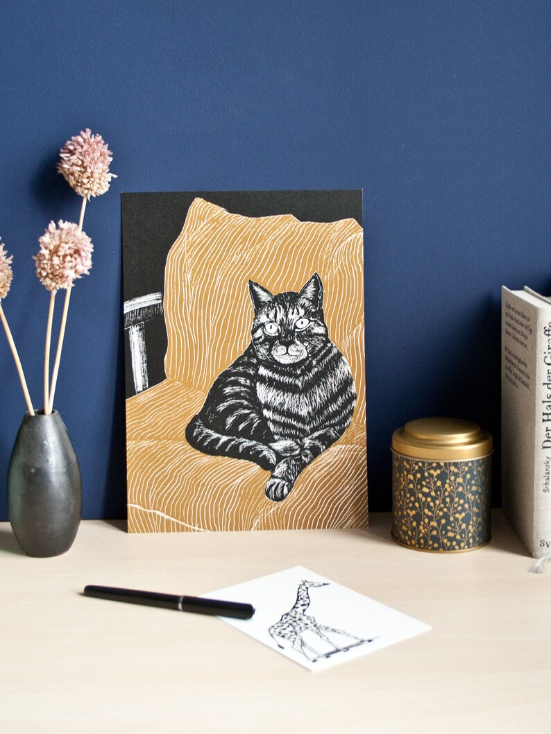 May include: A black and white cat art print on a black background, set against a gold-colored chair illustration. The print is displayed on a light-colored surface, with a black pen and a giraffe sketch. A black vase with dried flowers and a decorative tin are also present.