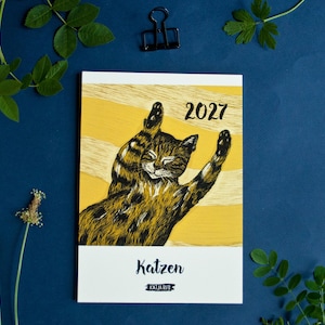 May include: A 2027 calendar featuring a yellow background with a black and brown illustrated cat stretching its paws upward. The text 2027 is printed in black at the top right, while the word Katzen and the name Katja Rub appear at the bottom.
