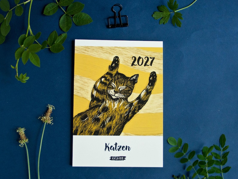 May include: A 2027 calendar featuring a yellow background with a black and brown illustrated cat stretching its paws upward. The text 2027 is printed in black at the top right, while the word Katzen and the name Katja Rub appear at the bottom.