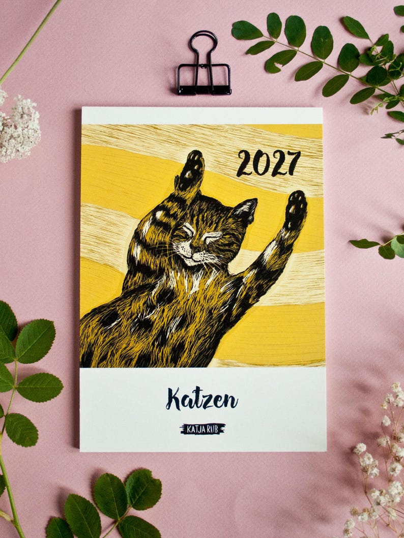 May include: A 2027 wall calendar featuring a black and brown tabby cat stretching on a yellow background with white horizontal stripes. The text "2027" appears in the upper right, while the word "Katzen" and "KATJA RUB" are printed at the bottom.