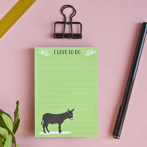May include: A light green notepad with the text "I LOVE TO DO" and a black donkey illustration. A black pen, a black binder clip, and gold tape are on a pink background. Green leaves are in the lower left corner. The notepad is for to-do lists.
