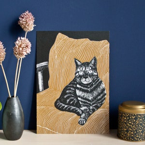May include: A black and white cat art print. The cat is sitting on a gold-colored chair with a black background. The print is displayed next to a black vase with dried flowers and a decorative tin.