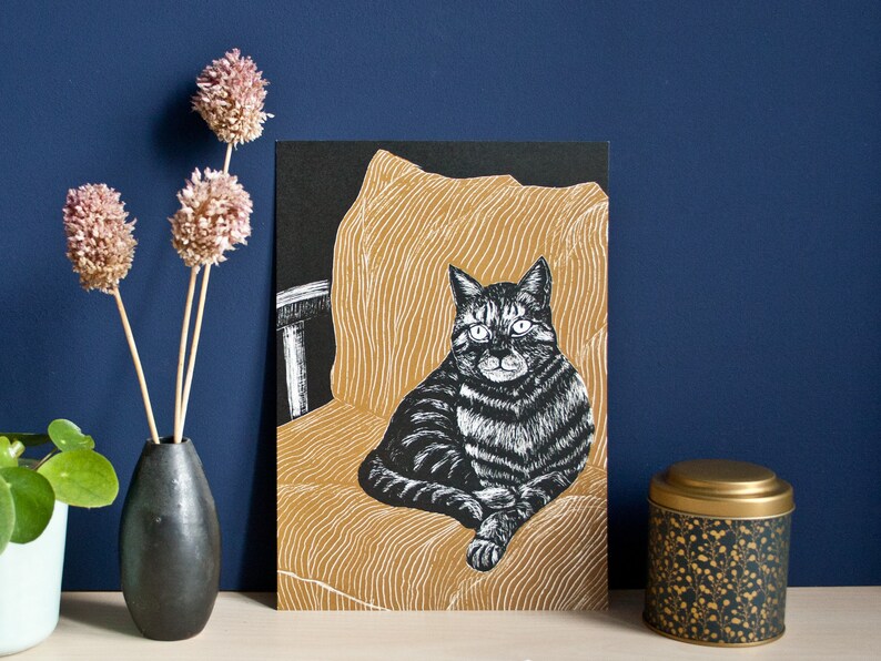 May include: A black and white cat art print. The cat is sitting on a gold-colored chair with a black background. The print is displayed next to a black vase with dried flowers and a decorative tin.