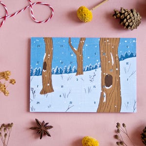 May include: A winter-themed advent calendar featuring a snowy forest scene with numbered boxes. The illustration shows brown tree trunks, white snow, and a blue sky. The calendar is surrounded by decorative elements on a pink background.