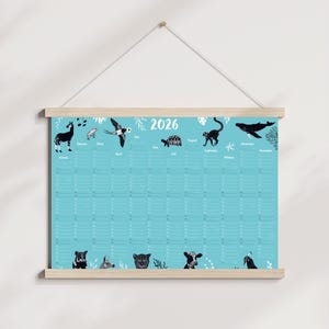 Könnte beinhalten: Ein Kalender für 2026 mit hellblauem Hintergrund und Holzrahmen. Der Kalender zeigt Illustrationen verschiedener Tiere, darunter ein Zebra, ein Vogel und ein Wal. Die Monate sind oben aufgeführt.