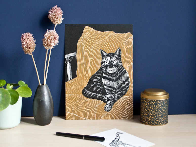 May include: A black and white cat illustration on a card, seated on a gold-colored chair. The card is propped up on a desk with a black vase holding dried flowers, a small tin, and a pen. The background is a dark blue wall.