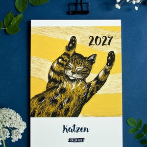 May include: A 2027 calendar featuring a black and brown tabby cat illustration with its paws raised against a yellow background with horizontal stripes. The text 2027 is printed in black, with the word Katzen and the name Katja Rub below.