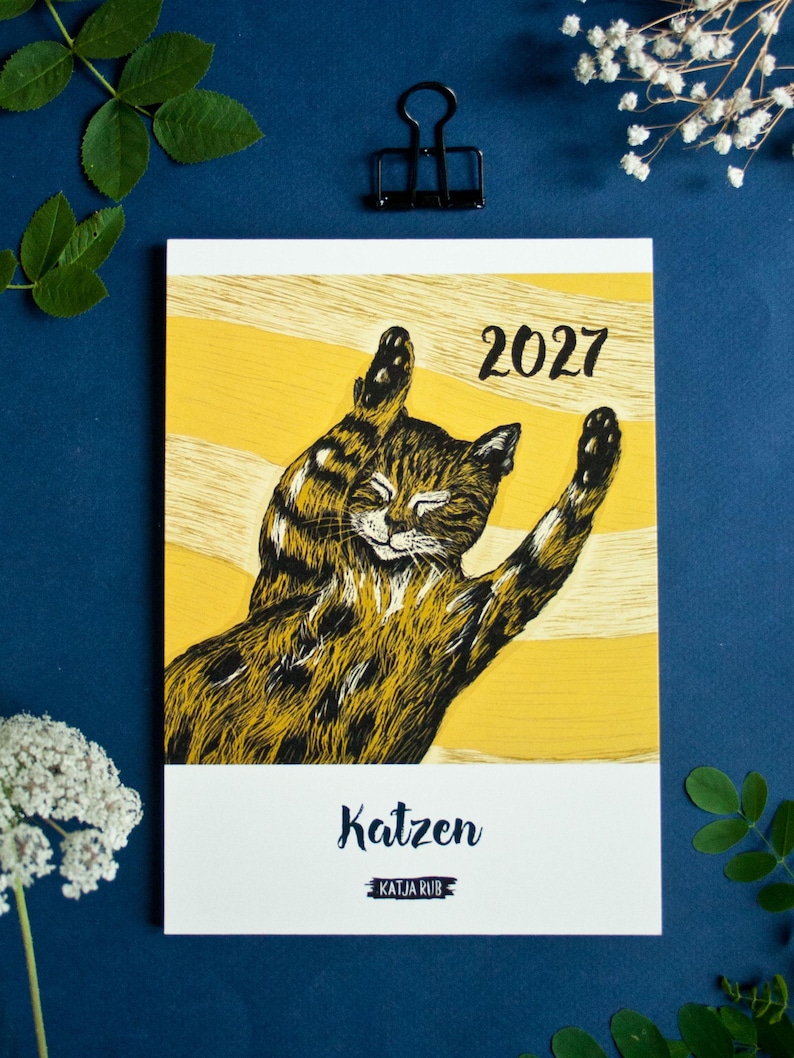 May include: A 2027 calendar featuring a black and brown tabby cat illustration with its paws raised against a yellow background with horizontal stripes. The text 2027 is printed in black, with the word Katzen and the name Katja Rub below.