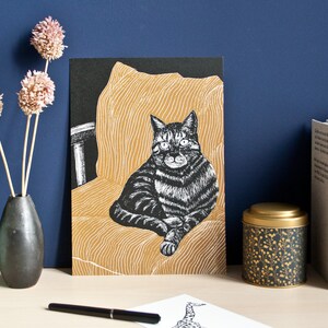 May include: A black and white cat art print. The cat is sitting on a gold-colored chair. The print is displayed on a desk with a vase of dried flowers, a small plant, and a decorative tin.
