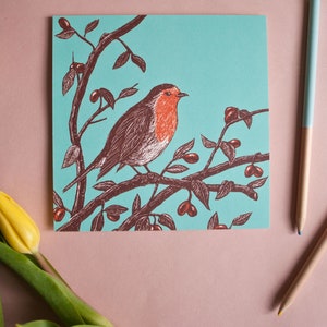 May include: A greeting card with a robin perched on a branch with red berries. The robin is brown with a red breast and is set against a turquoise background.
