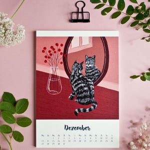 May include: A wall calendar page for the month of Dezember featuring a black and white striped cat sitting in front of a round mirror. The illustration includes a clear glass vase with red berries on a dark red surface against a pink background.