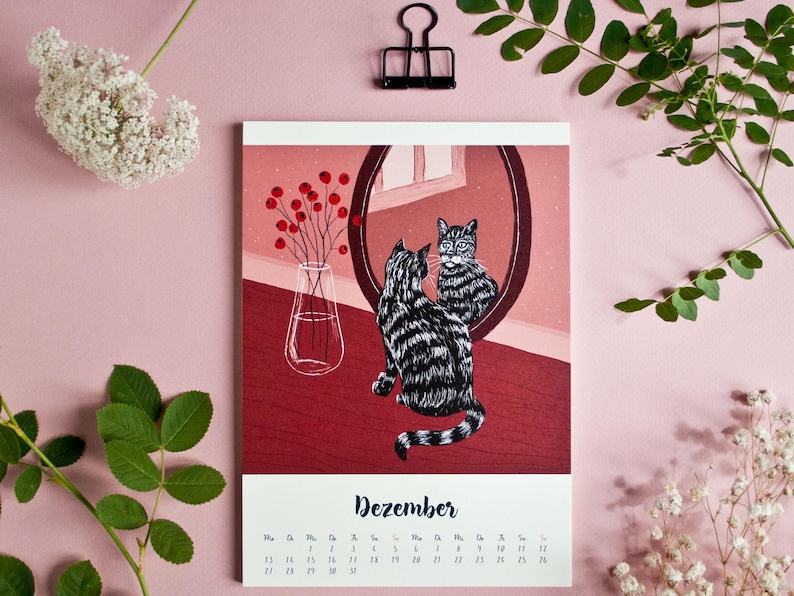 May include: A wall calendar page for the month of Dezember featuring a black and white striped cat sitting in front of a round mirror. The illustration includes a clear glass vase with red berries on a dark red surface against a pink background.
