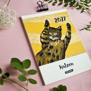 May include: A 2027 calendar featuring a black and brown tabby cat illustration with raised paws against a yellow and white striped background. The cover displays the text 'Katzen' and 'KATJA RUB' in black lettering on a white lower section.
