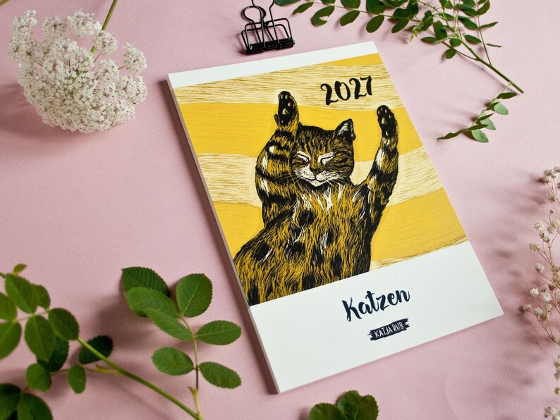 May include: A 2027 calendar featuring a black and brown tabby cat illustration with raised paws against a yellow and white striped background. The cover displays the text 'Katzen' and 'KATJA RUB' in black lettering on a white lower section.