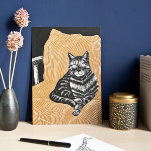 May include: A black and white cat art print. The cat is seated on a yellow chair with a black background. The print is displayed on a wooden surface with a vase of dried flowers, books, and a decorative tin.