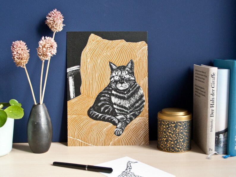 May include: A black and white cat art print. The cat is seated on a yellow chair with a black background. The print is displayed on a wooden surface with a vase of dried flowers, books, and a decorative tin.