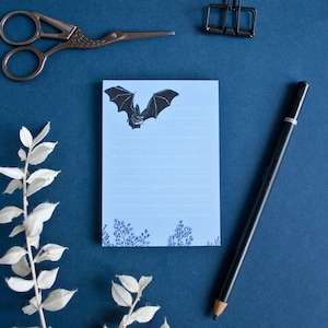 Op de afbeelding: Een blauwe notitieblok met een zwart vleermuis silhouet en gestreept papier. De notitieblok ligt op een blauw oppervlak met een witte tak en een zwart potlood.