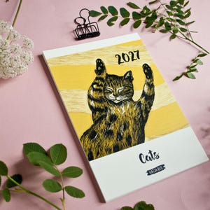 May include: A 2027 calendar featuring a black and brown tabby cat illustration with raised paws against a yellow and white striped background. The cover includes the text 2027, Cats, and Katja Rub in black lettering on a white rectangular base.