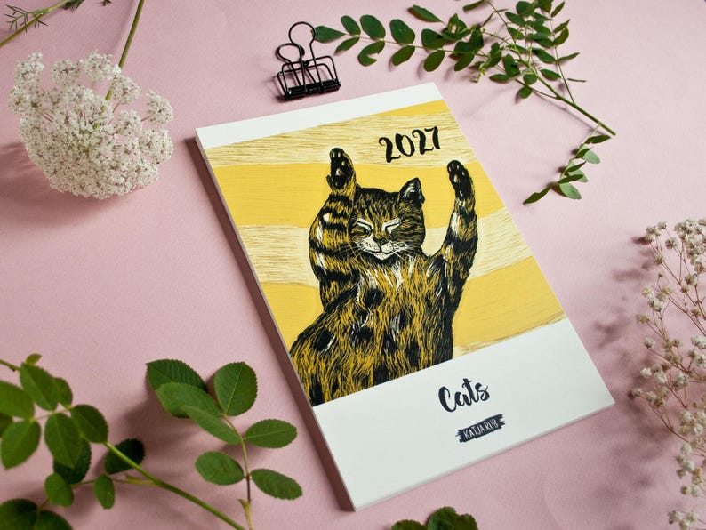 May include: A 2027 calendar featuring a black and brown tabby cat illustration with raised paws against a yellow and white striped background. The cover includes the text 2027, Cats, and Katja Rub in black lettering on a white rectangular base.
