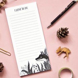 May include: A white notepad with black lines and the text "I LOVE TO DO" at the top. The notepad has a black and white illustration of a fox in a forest on the bottom. The notepad is on a pink surface with other decorative items.