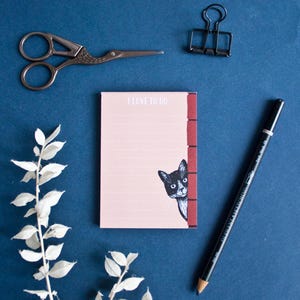 Può includere: Un blocco note rosa con il testo "I love to do" stampato sulla parte anteriore. Un gatto bianco e nero spunta da dietro un muro di mattoni rossi sul lato destro del blocco note.