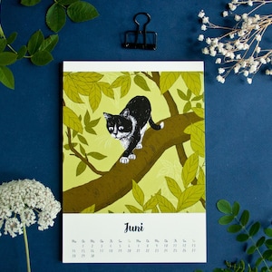 May include: A wall calendar page for the month of June featuring a black and white cat walking along a brown tree branch surrounded by light green leaves. The text "Juni" is printed in black above the calendar grid for the month.