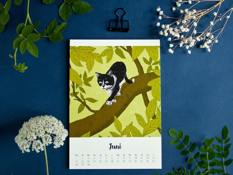 May include: A wall calendar page for the month of June featuring a black and white cat walking along a brown tree branch surrounded by light green leaves. The text "Juni" is printed in black above the calendar grid for the month.