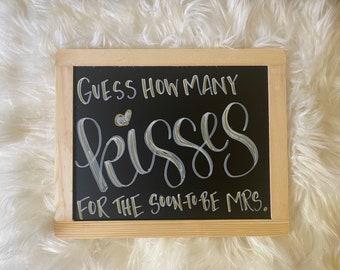 May include: A black chalkboard with a wooden frame, featuring white and gold lettering that reads "Guess how many kisses for the soon-to-be Mrs."