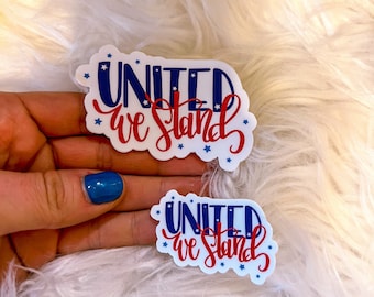 May include: Two white stickers with the text "UNITED we STAND" in red and blue. The word "UNITED" is in blue block letters with white stars, and "we STAND" is in red script. The stickers are on a fluffy white surface.