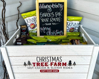 May include: A white wooden crate with the text "Christmas Tree Farm" and "Hot Cocoa & Sleigh Rides" printed on it. The crate is filled with bags of snacks and a chalkboard sign that reads "Delivery Drivers Snacks for Your Route - Thank You - Merry Christmas".