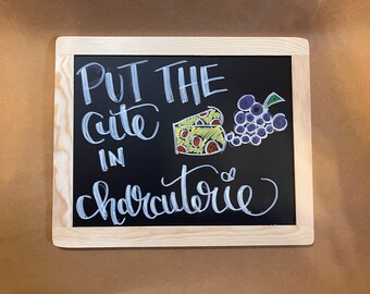 May include: A black chalkboard with a wooden frame, featuring white chalk lettering that reads "Put the cute in charcuterie". The chalkboard also includes a drawing of a wedge of cheese and a bunch of grapes.